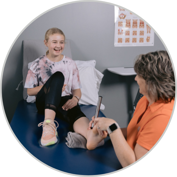A cheerful young girl in casual attire sits on a medical table, smiling as a healthcare professional consults her foot. The setting is bright and relaxed.